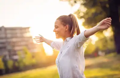 An image of a woman standing outside in nature with her eyes closed and arms raised.