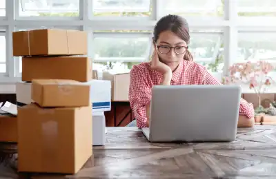 Small business owner on their laptop with boxes of deliveries next to them.