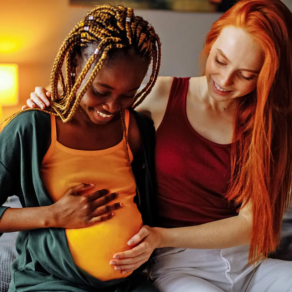 Two women sat together. One is pregnant and the other is embracing her and holding her abdomen