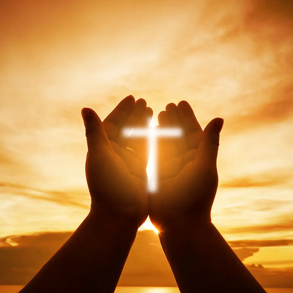 Hands open palm up in worship in front of a rising sun. The sign of the cross is glowing on the hands