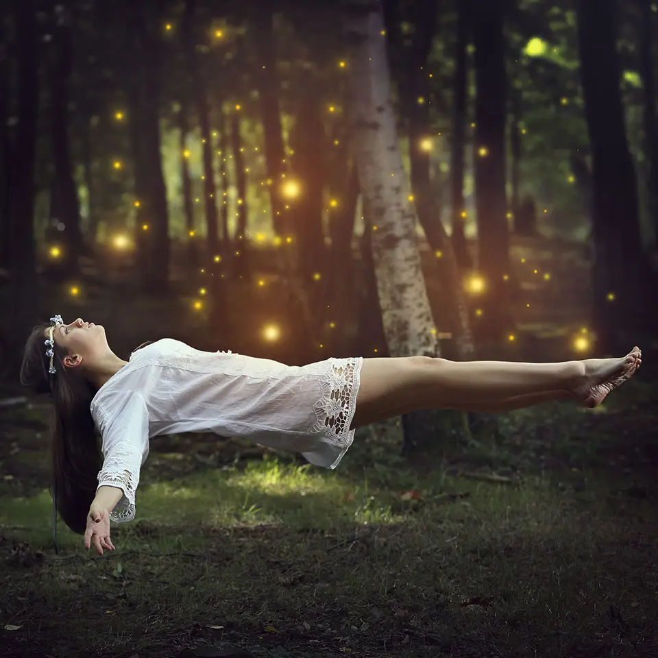 A woman lying down but floating above the ground in a forest of trees and glowing lights