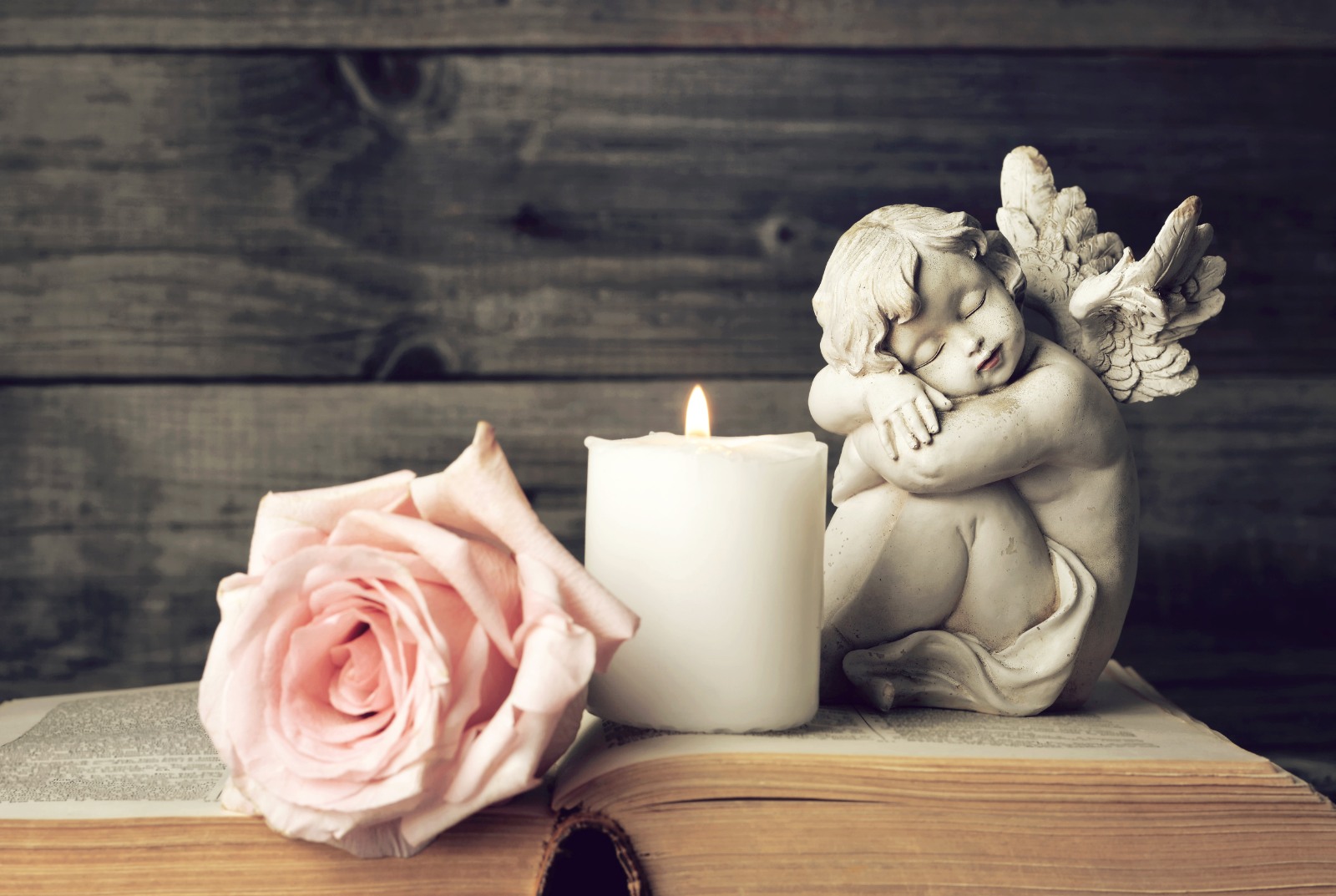 A statue of an angel next to a candle and a pink rose, on top of a book.
