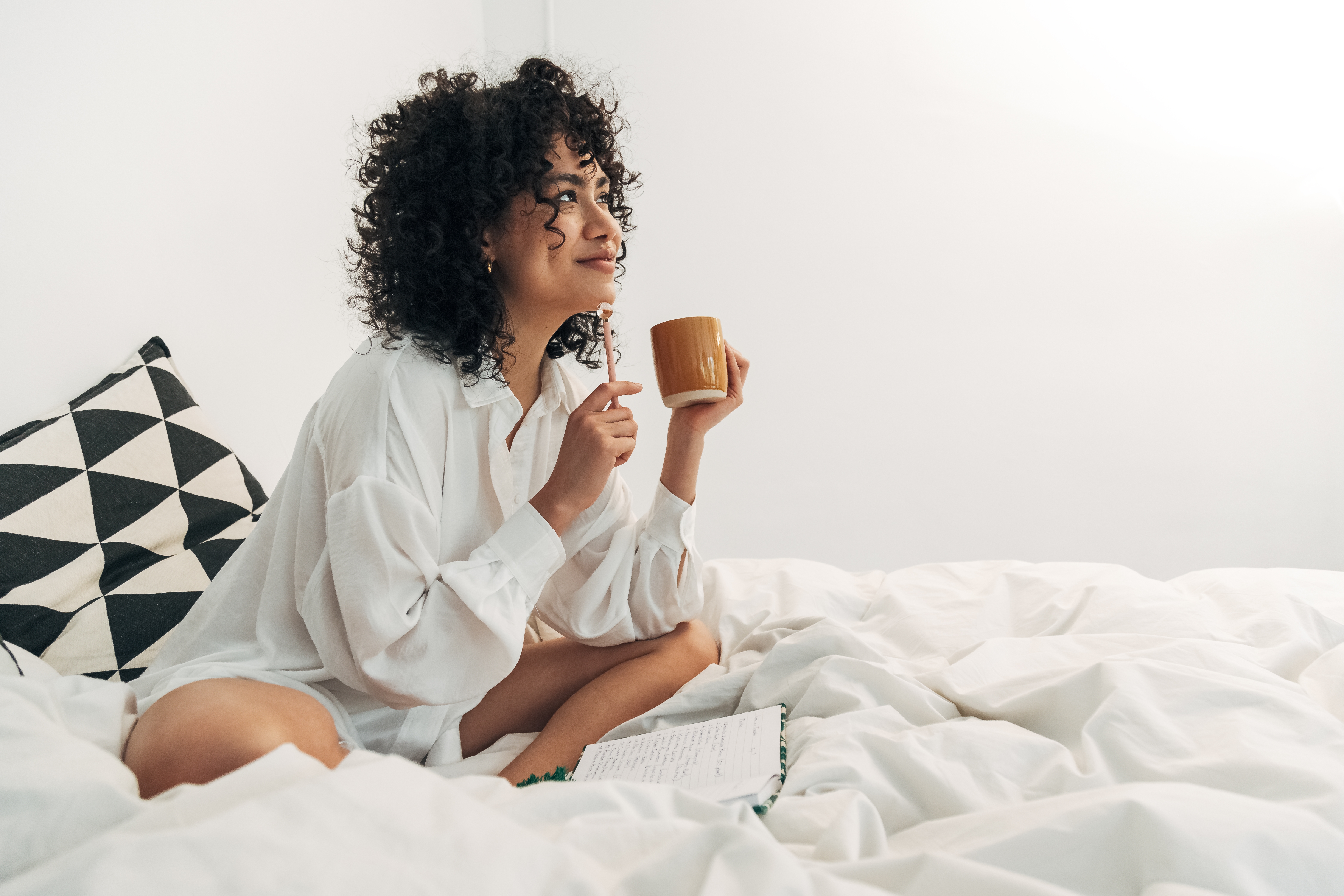 Happy woman in bed journaling with notebook, pen and cup of coffee