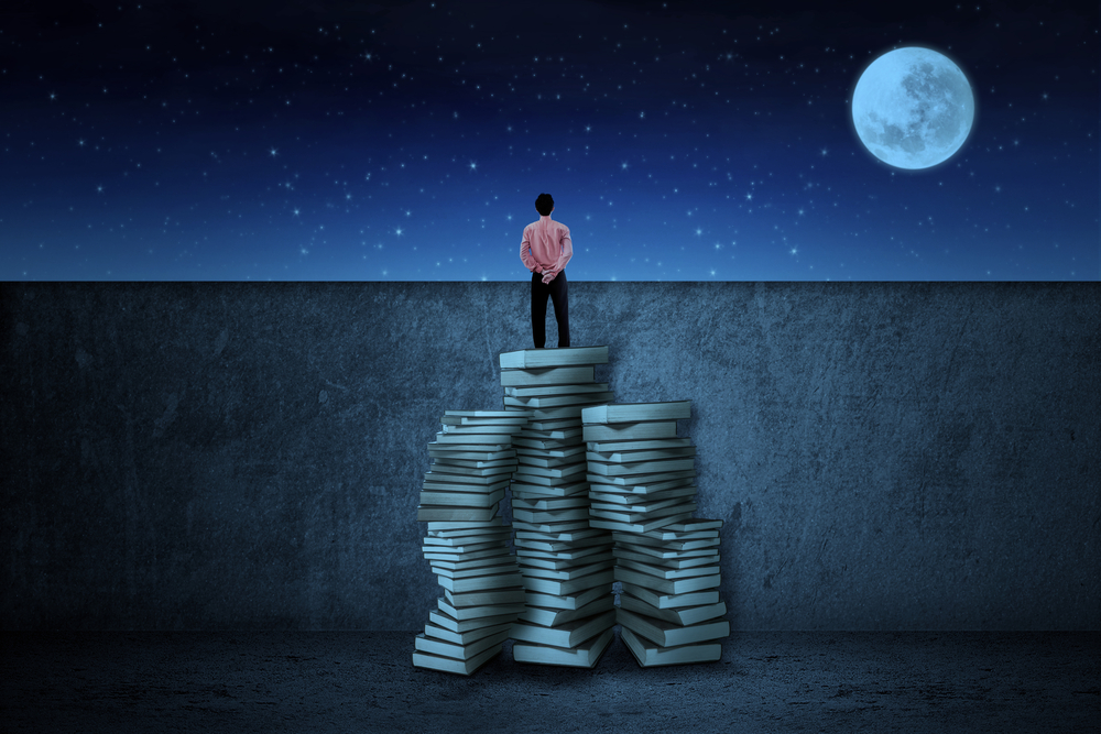 Businessman standing on top of a pile of books looking at a full moon
