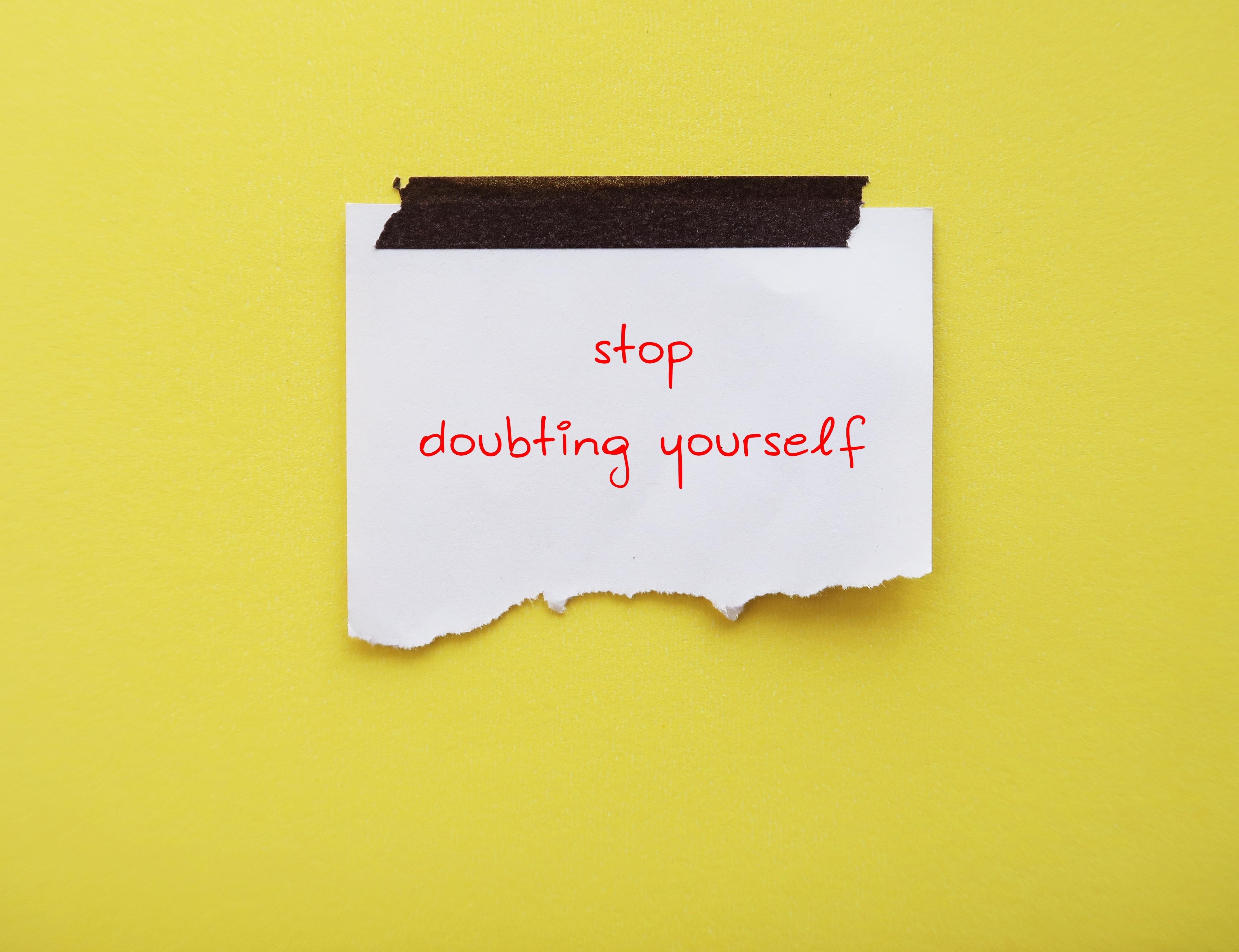 A white piece of paper on a yellow background with 'stop doubting yourself' written on it