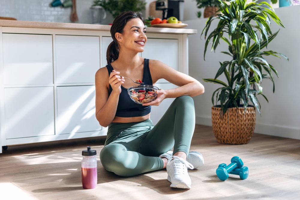 A smiling woman sat eating a healthy bowl of food with weights and a smoothie next to her