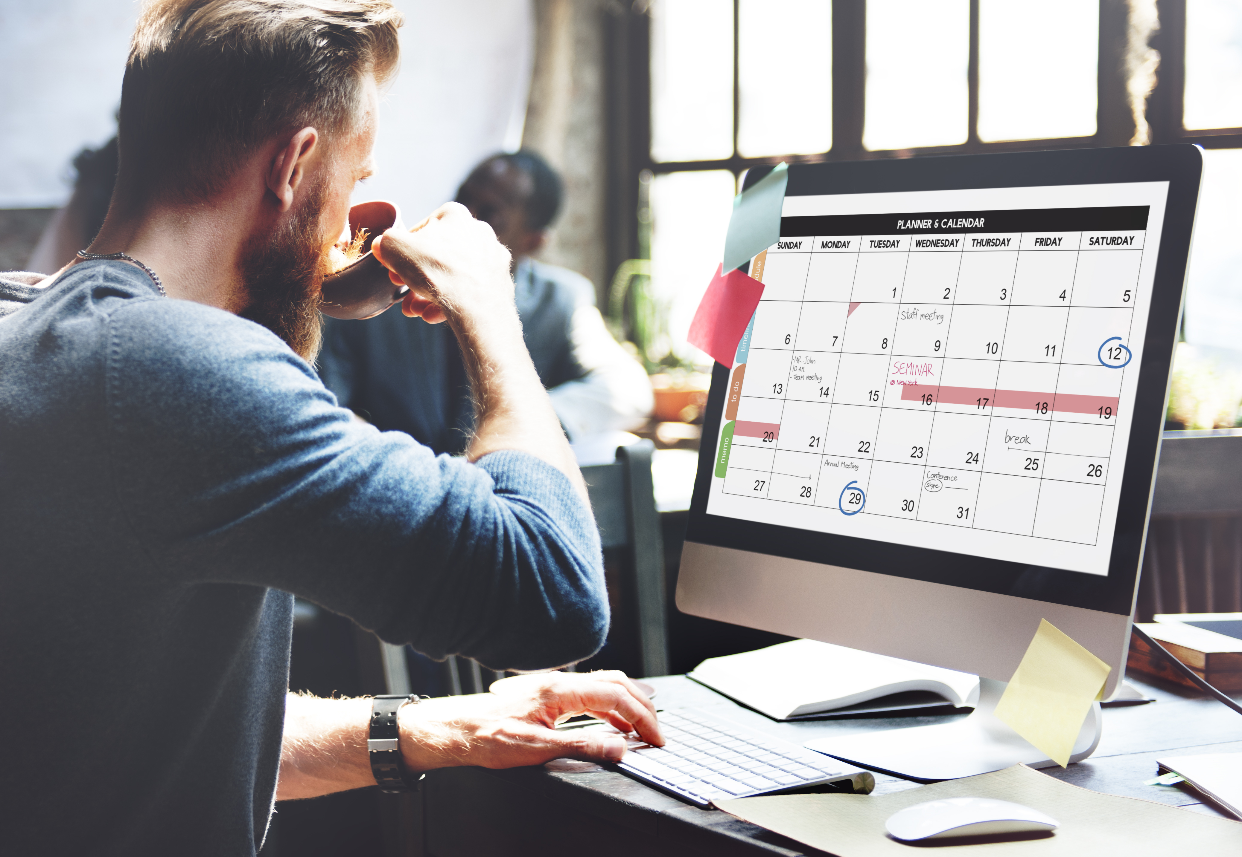 A man sat at a computer looking at a calendar and drinking a hot drink from a mug