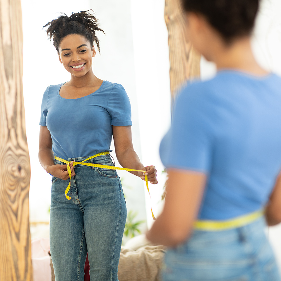 Happy woman measuring her waist with tape standing in front of a mirror