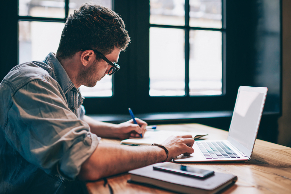 A young man with a laptop writing in a notepad