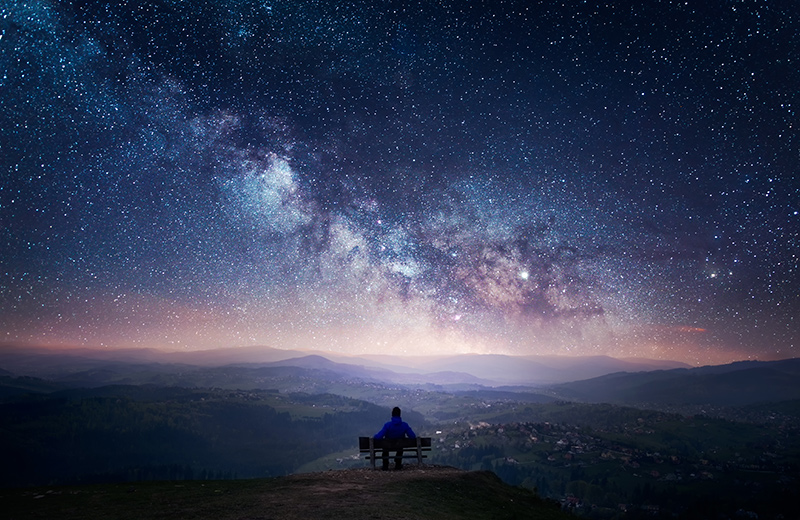 A man sitting on a bench staring at a starry sky with a Milky Way and a mountain landscape