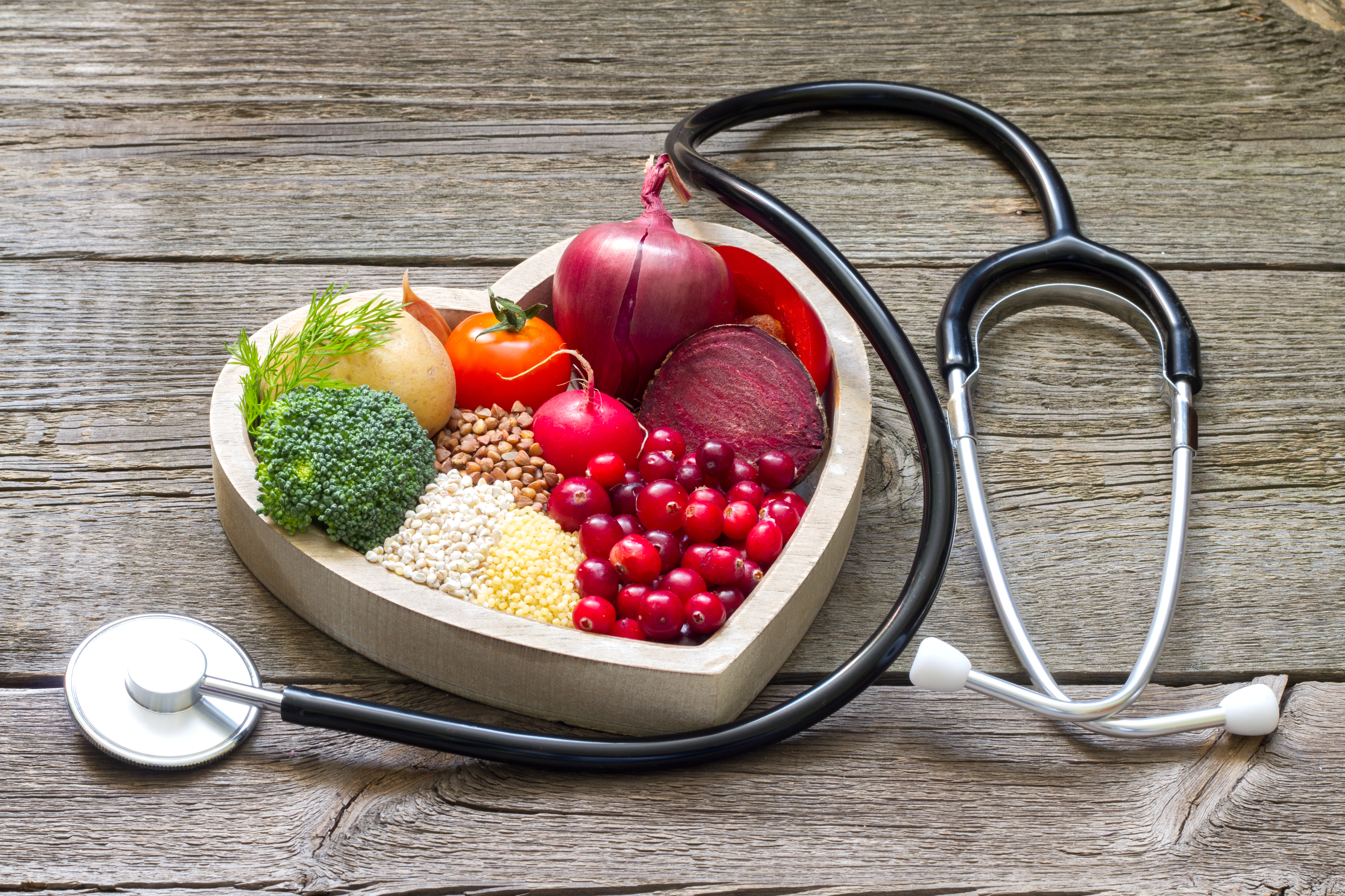 A heart shaped bowl filled with various fruits and vegetables and a doctors stethoscope next to it 