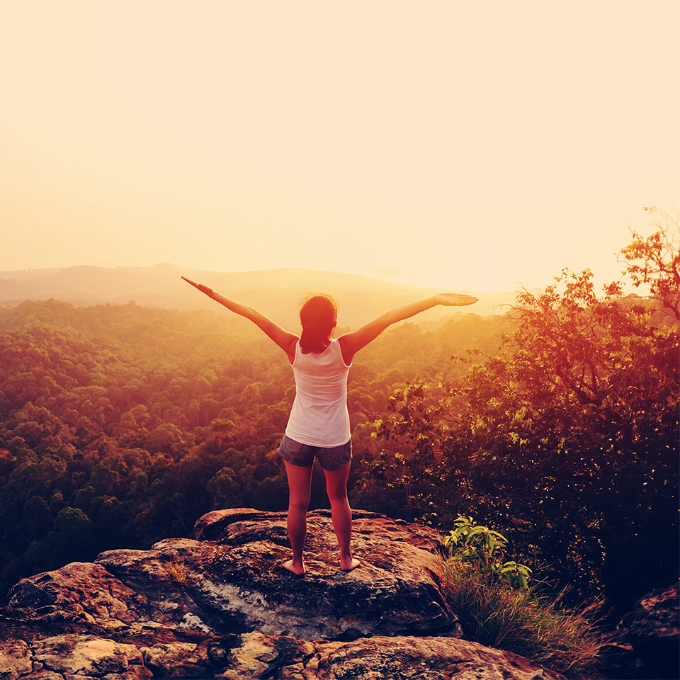 A person on top of a mountain with their hands up and outstretched in celebration, looking towards the rising sun
