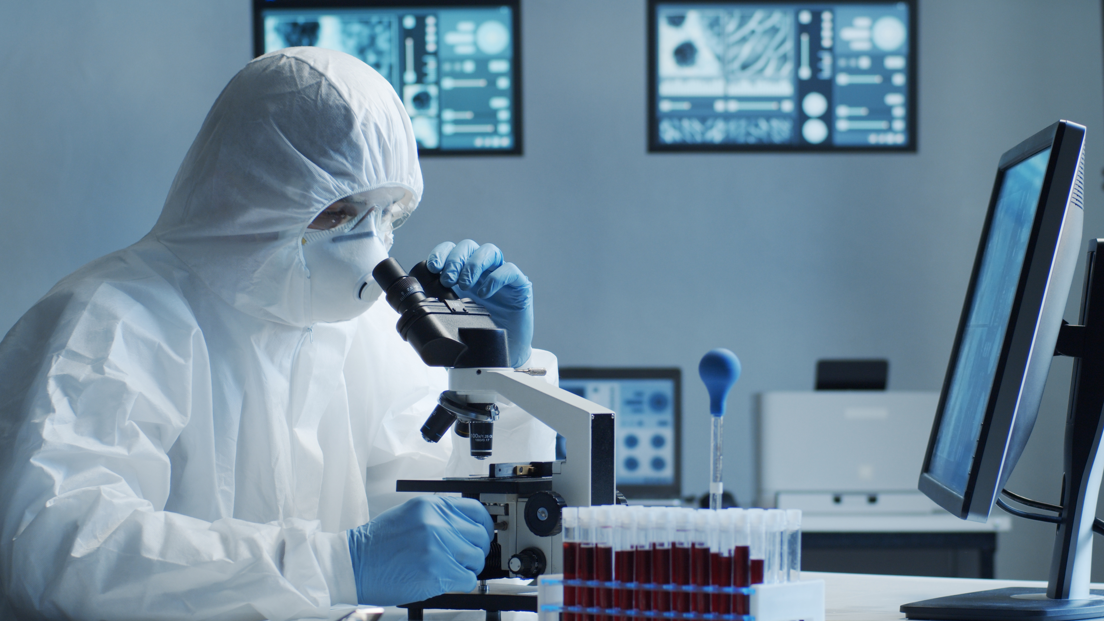 A scientist in scrubs, a face mask and goggles looking through a microscope