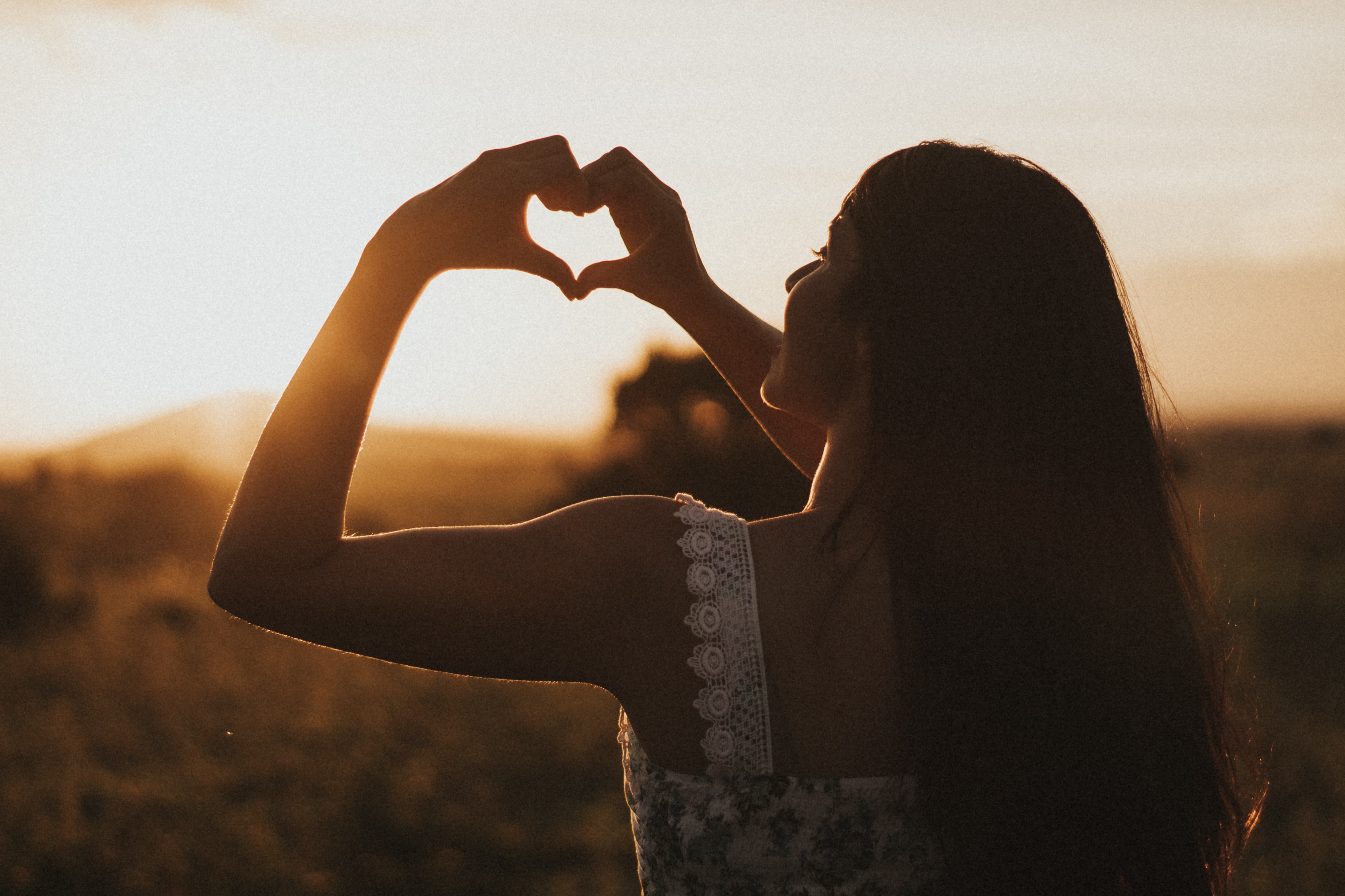 A woman making a heart shape against a sunset sky.