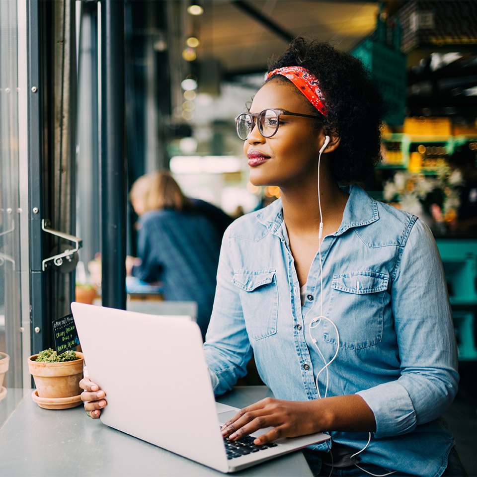 Smiling person looking through a window while sitting in a café working on a laptop.