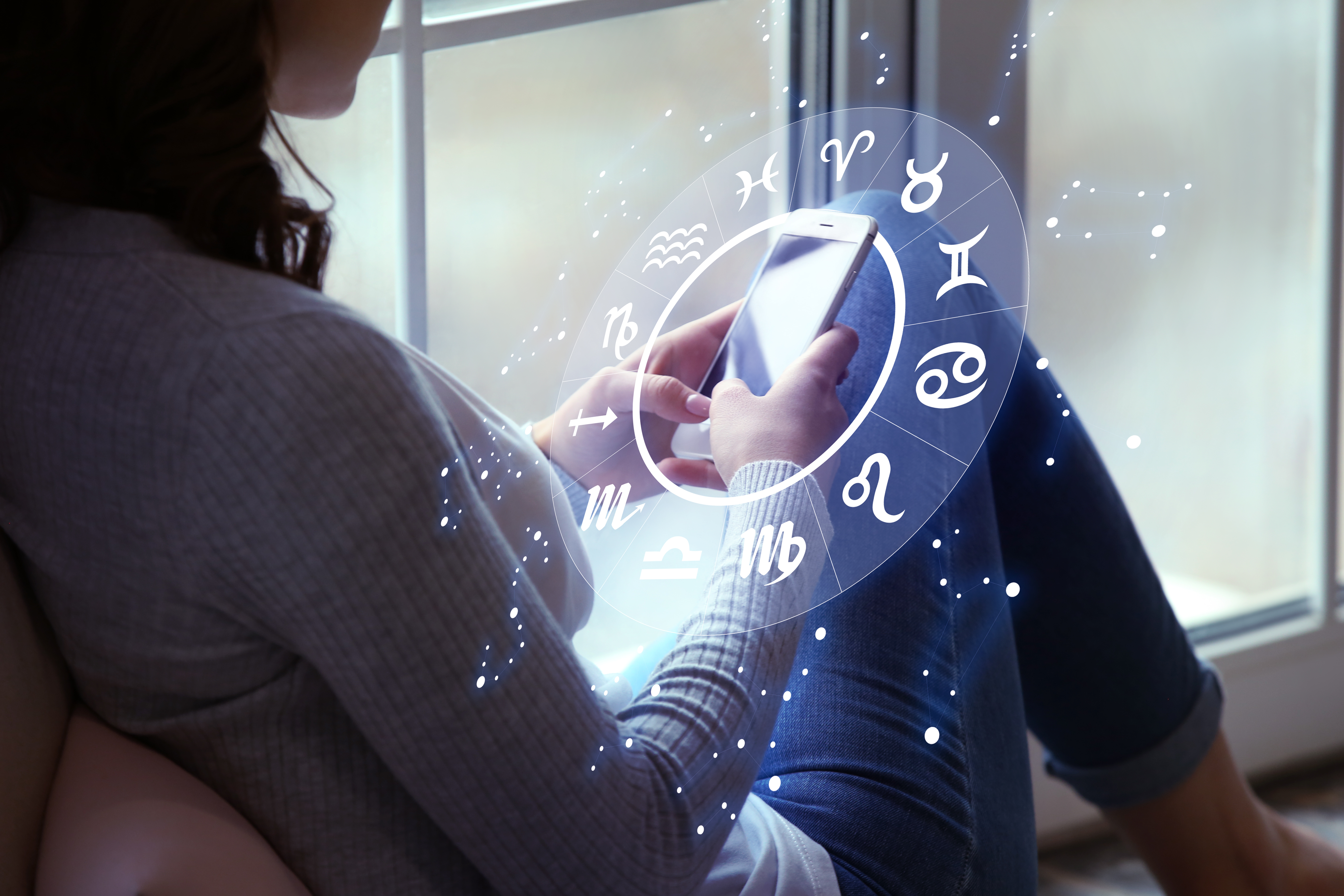 A woman sat next to a window looking at her phone screen with a zodiac wheel around the phone