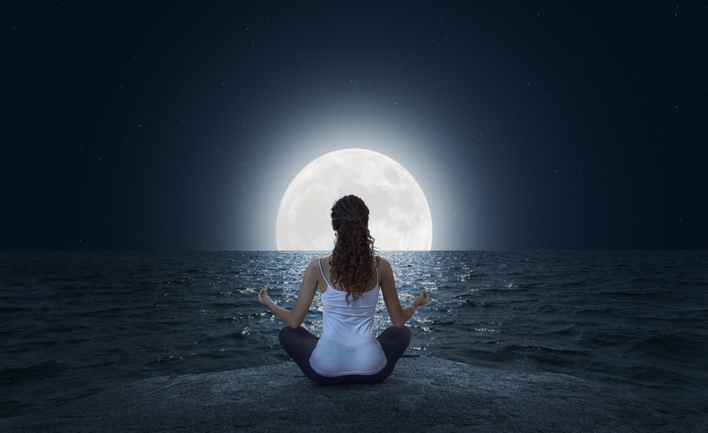 A woman meditating next to he sea in front of a full moon