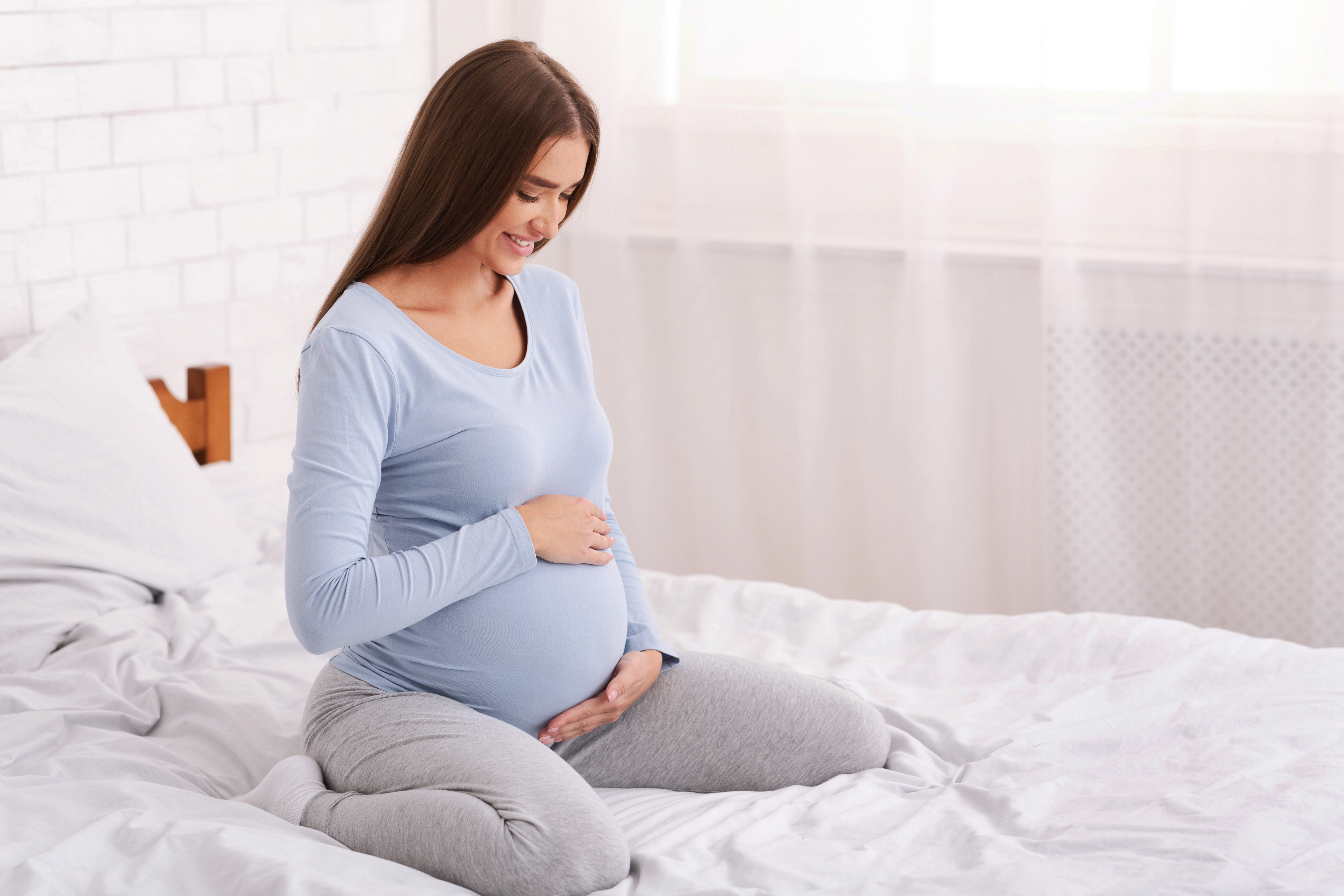 Pregnant lady cradling bump on bed in a white room