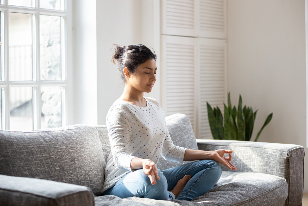 Woman sat cross legged on a sofa with her eyes closed, meditating