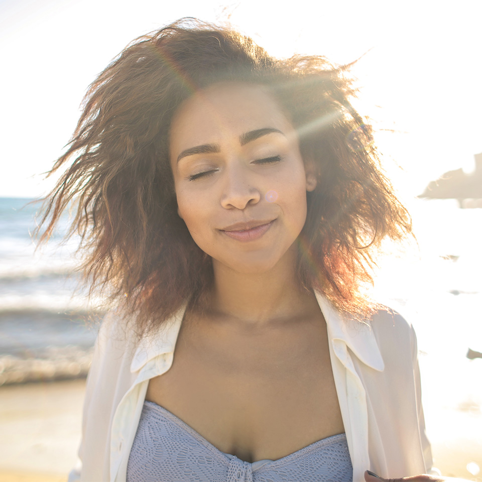 A woman with her eyes closed as she stands on a beach, practising rebirthing breathwork.