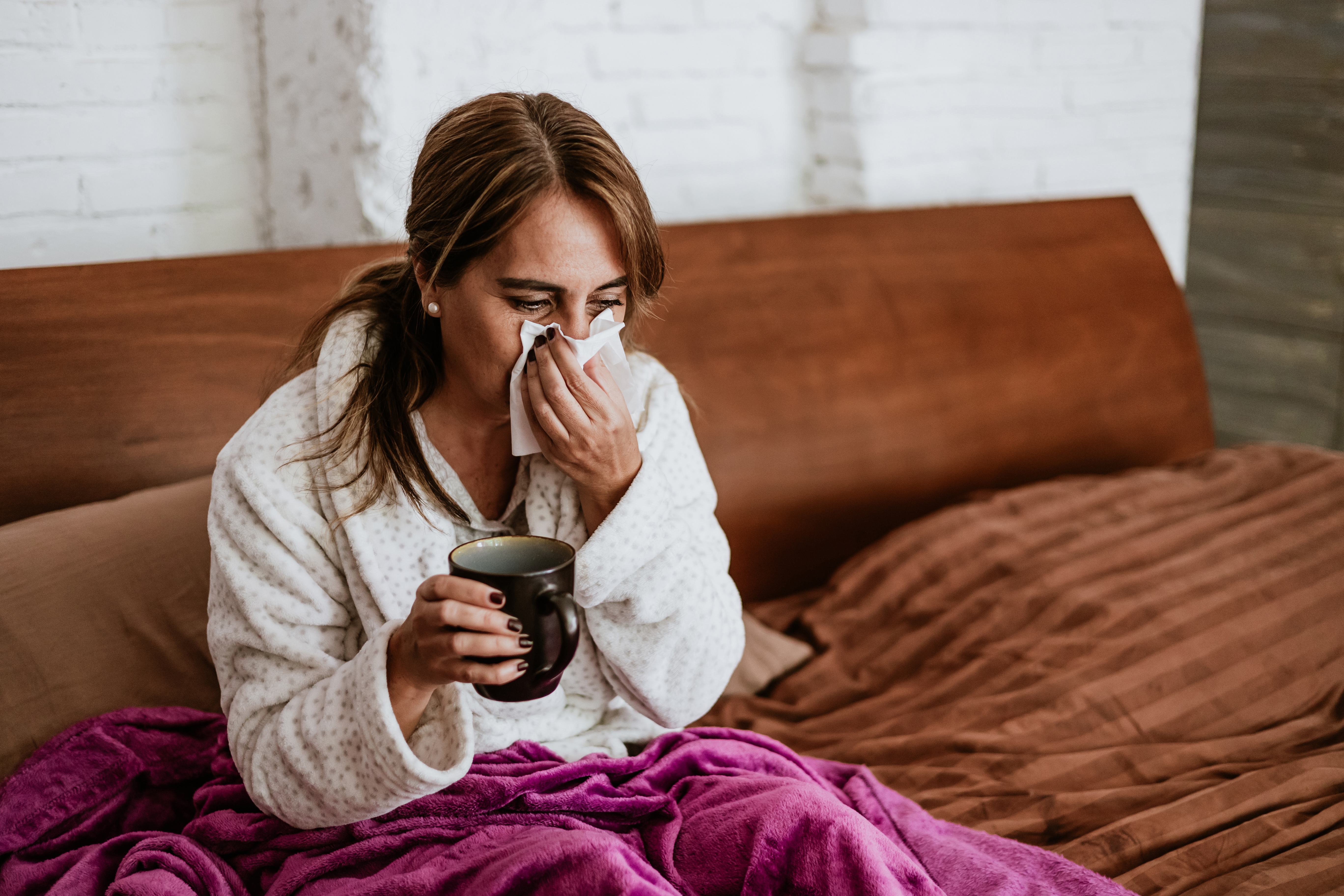 Woman ill with the flu blowing her nose in bed, with a cup of tea