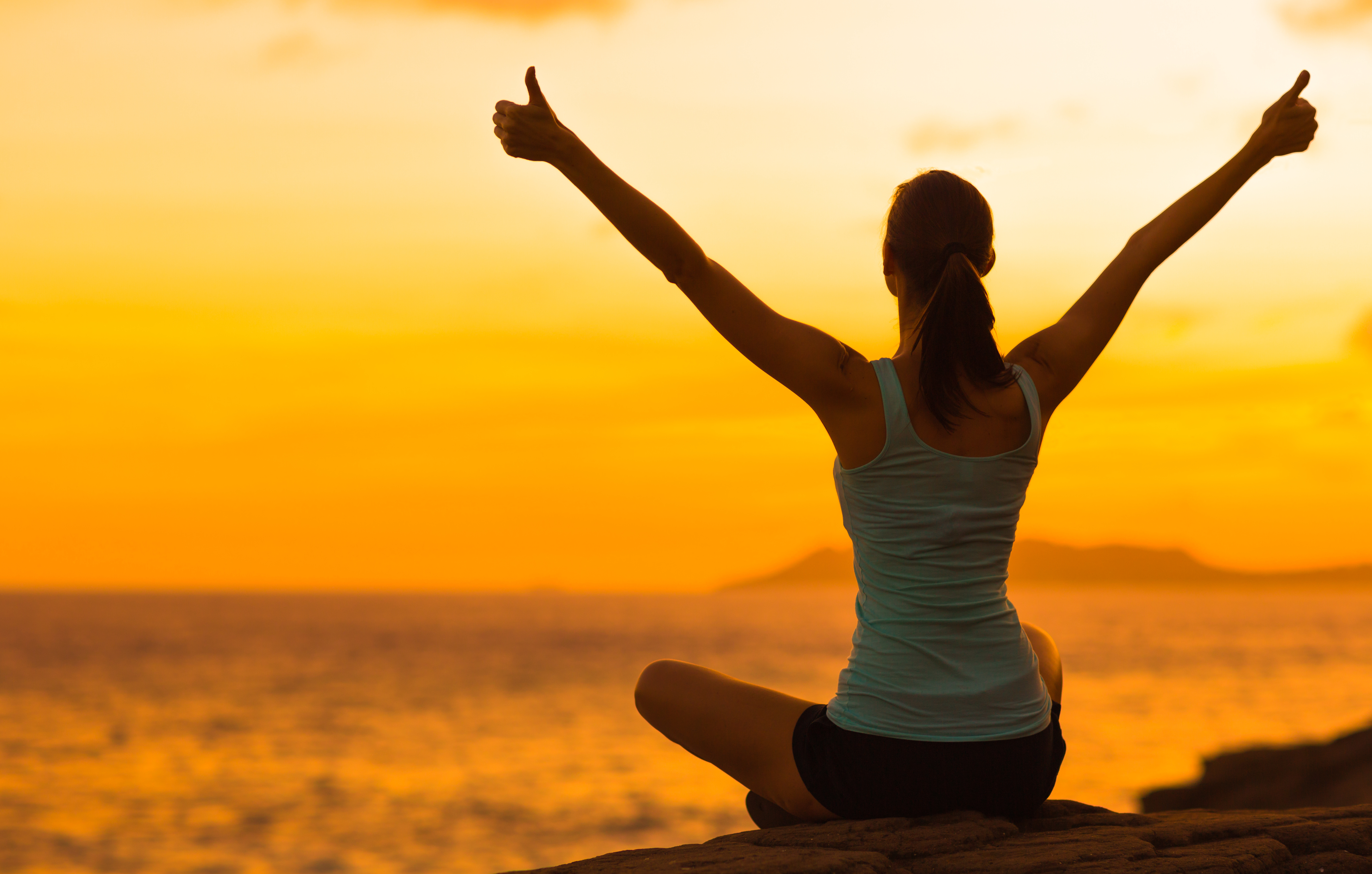 A woman sat on rocks next to the sea with her hands in the air and her thumbs up