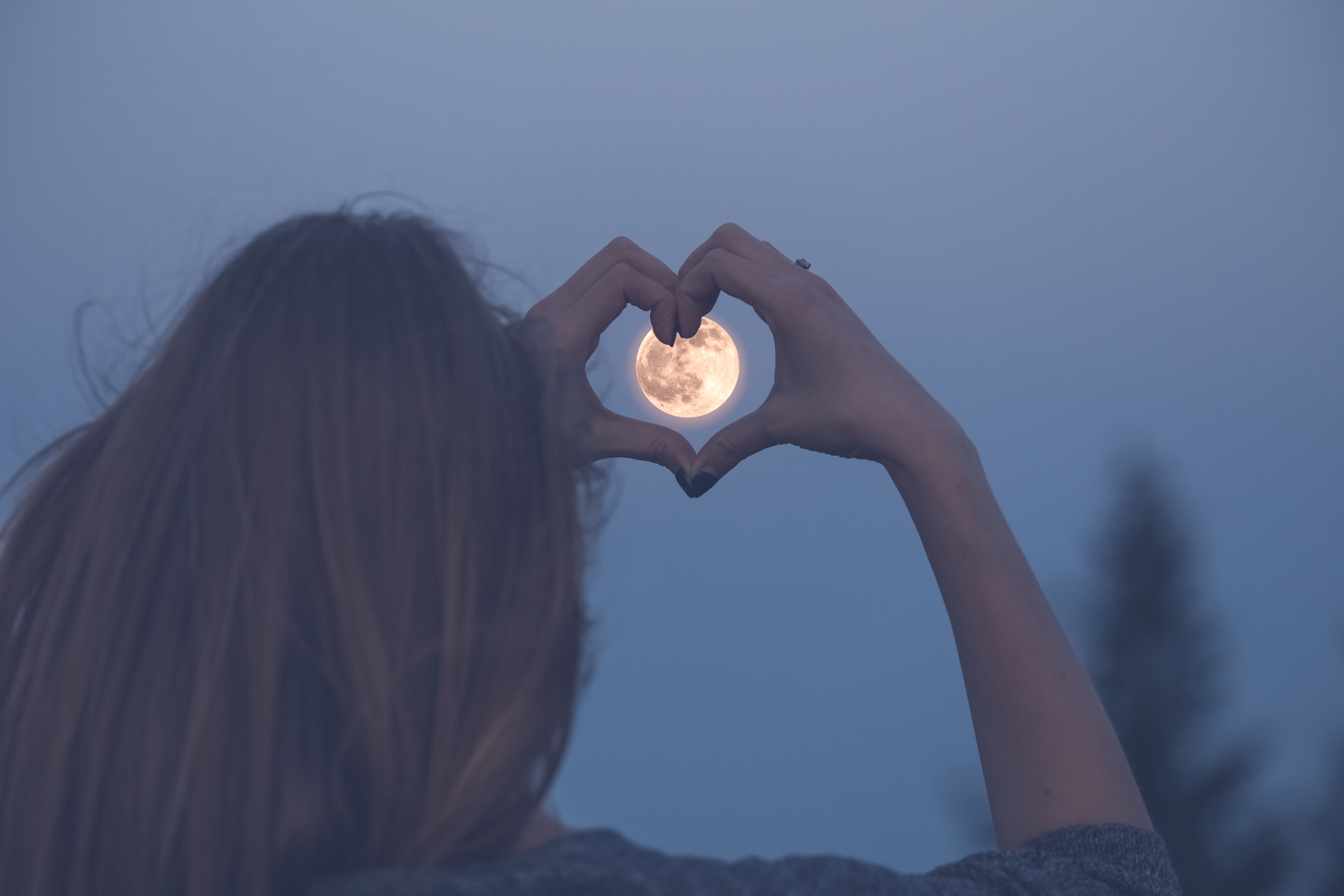 A woman making a heart shape with her hands over a full moon