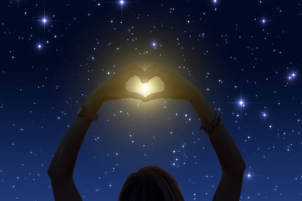 Silhouette of a person making a love heart with two hands, holding them up to a starry night sky with a bright star in the middle