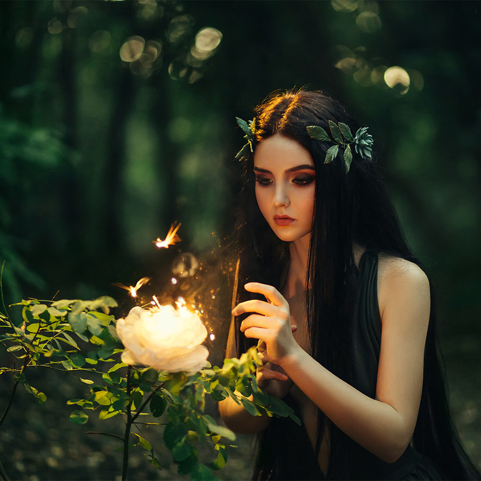 A fairy with long hair in a forest, looking at a flaming, fiery flower surrounded by butterflies.