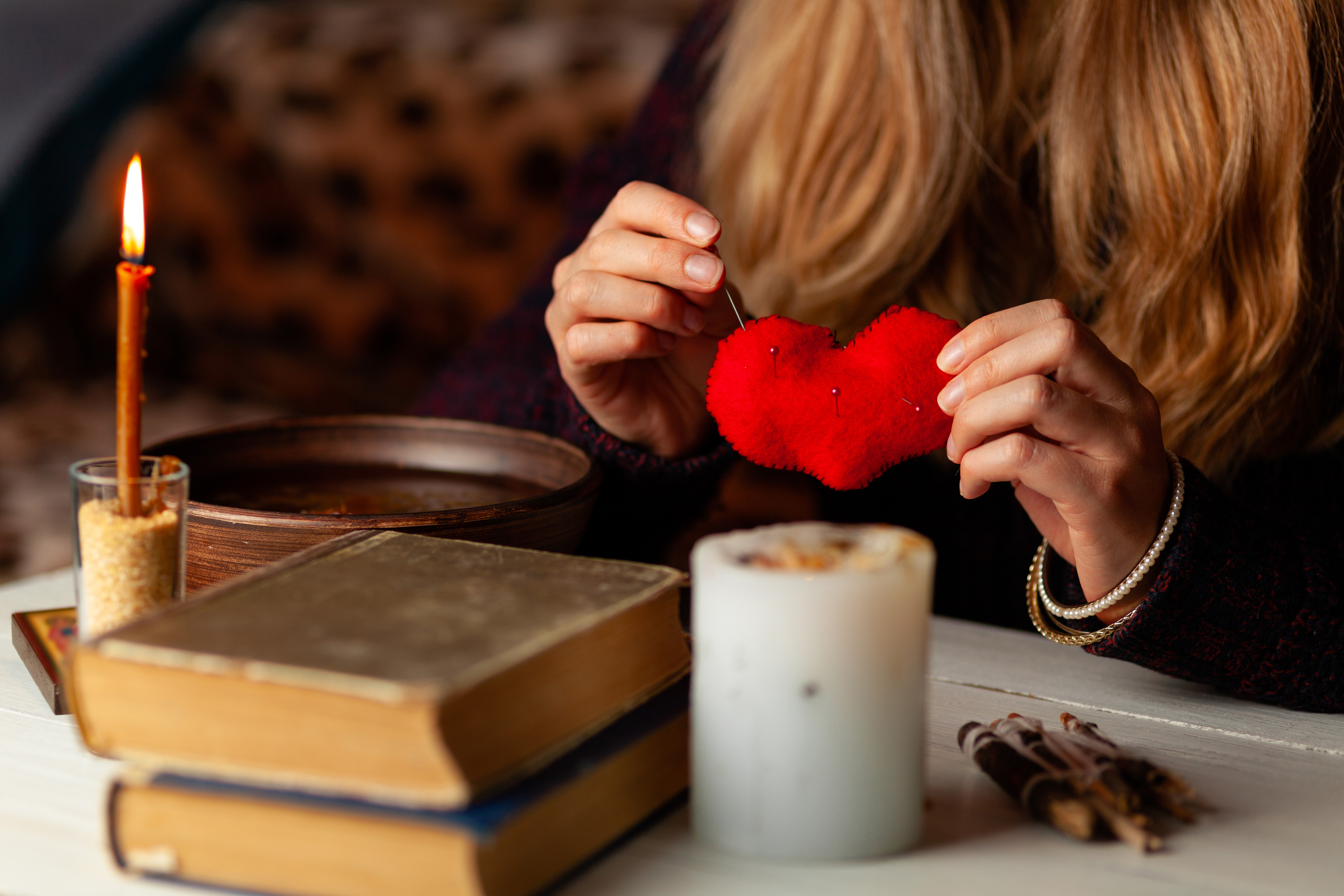 Someone sticking pins into a heart shaped pin cushion next to a lit candle