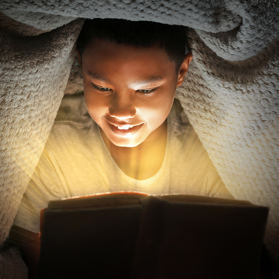 Child reading a book under blanket at night